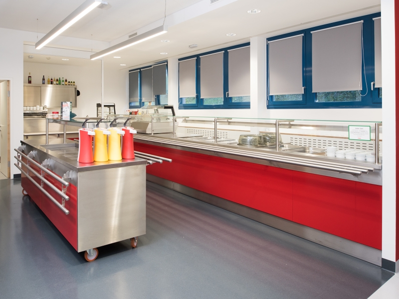 Speiseausgabe in Kantine: Tablettrutsche mit Rundrohren, Zugriff- und Spuckschutz aus Glas und fahrbares Salatbuffet; farbliche Akzente mit Feuerwehr-Rot