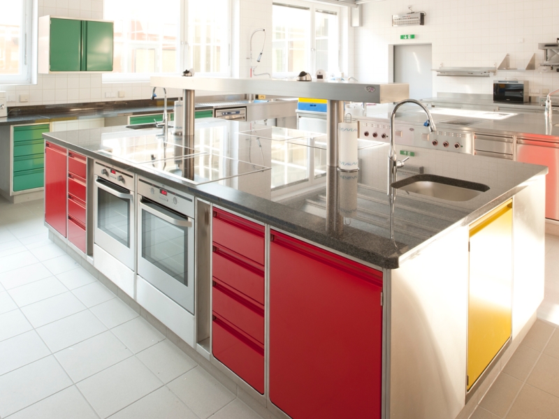 Schulküche in Edelstahl mit Farbleitsystem für einfaches Auffinden von Kochutensilien; Unterschränke in Hygiene H2; Abdeckungen in Edelstahl bzw. Stein